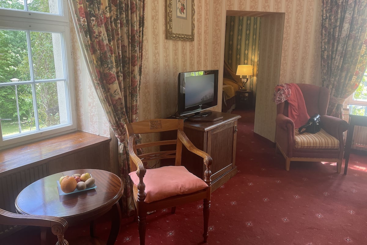Quarto com cadeira de madeira, uma mesinha com frutas, televisão sobre um suporte de madeira e piso coberto por um tapete vermelho.
