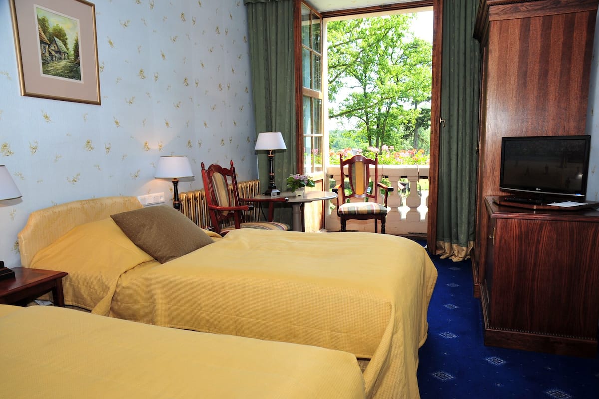 Quarto de hotel com duas camas, televisão, uma mesinha com abajur e vista para o verde lá fora.