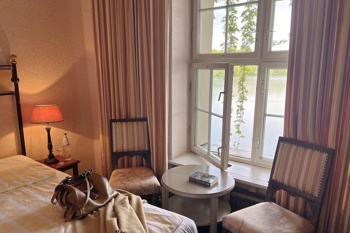 Quarto de hotel com uma cama, uma mesinha, duas cadeiras, um abajur e uma janela com vista para vegetação.
