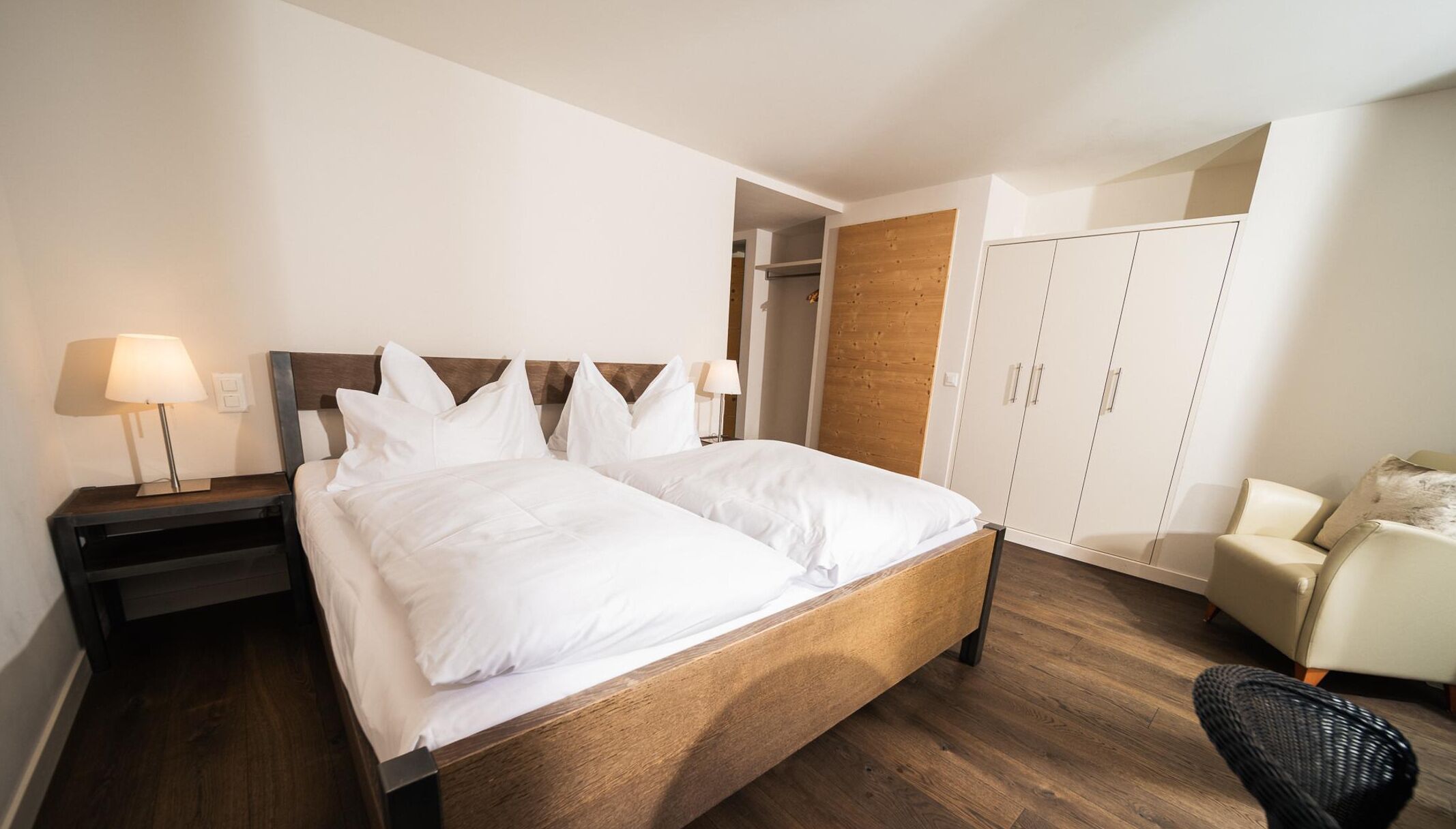 Una cama bien hecha con s ábanas blancas, un cabecero de madera, una mesita de noche con lámpara, una silla y un armario.