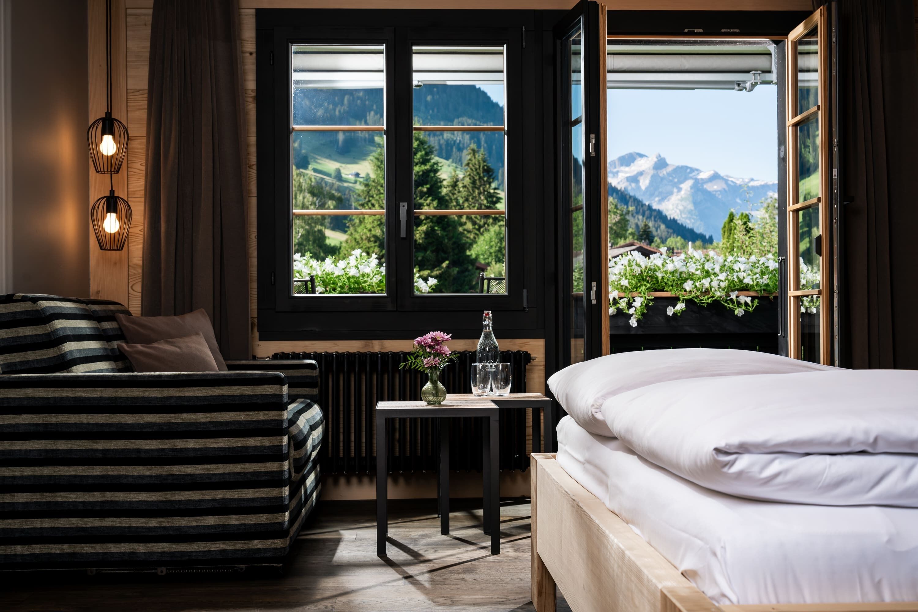 A bedroom with a bed, a sofa, a table with a vase of flowers, and a view of mountains through a large window.