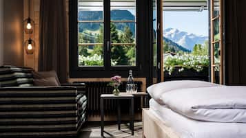 A bedroom with a bed, a sofa, a table with a vase of flowers, and a view of mountains through a large window.