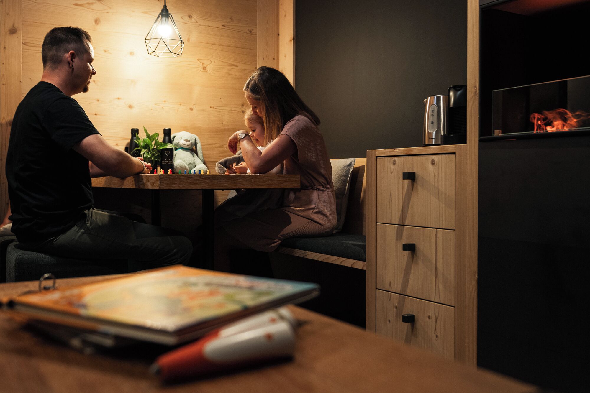 A wooden table with a laptop, a pen, and a book. There is a teddy bear and a bottle on the table.