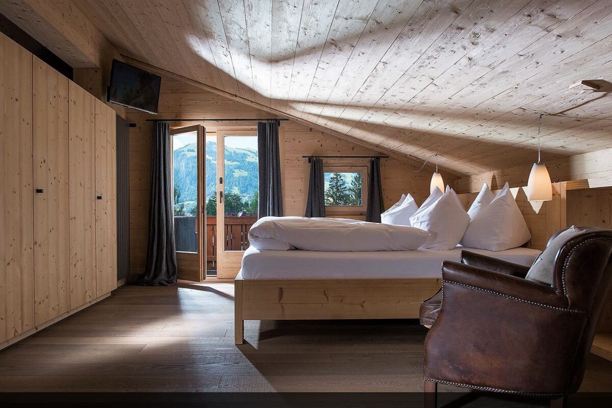 Una suite tipo chalet con interior de madera, una cama con ropa blanca, un sillón de cuero marrón y vistas a las montañas a través de las ventanas.