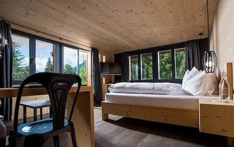 A room with a wooden bed, a desk, and a chair, large windows, and a view of trees.
