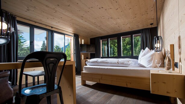 A room with a wooden bed, a desk, and a chair, large windows, and a view of trees. - Arc-en-Ciel Gstaad (Saanen)
