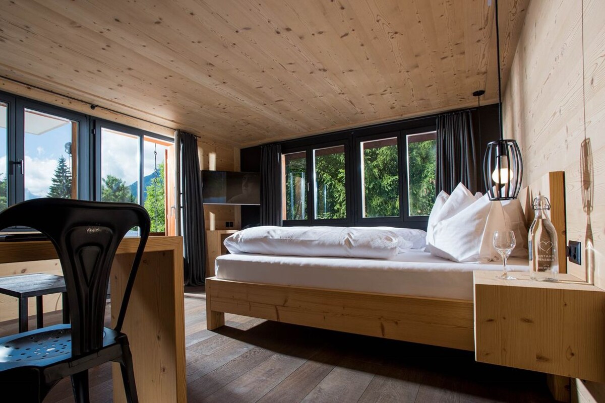 Una habitación con una cama de madera, un escritorio, una silla, ventanas amplias y vistas a árboles.