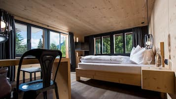 A room with a wooden bed, a desk, and a chair, large windows, and a view of trees.