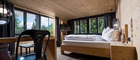 A room with a wooden bed, a desk, and a chair, large windows, and a view of trees.