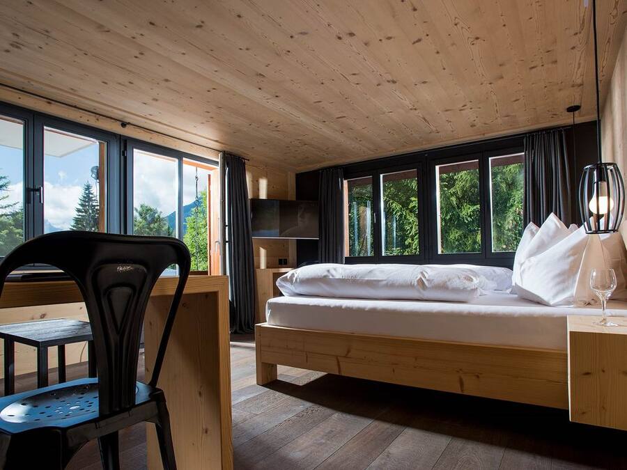 A room with a wooden bed, a desk, and a chair, large windows, and a view of trees.