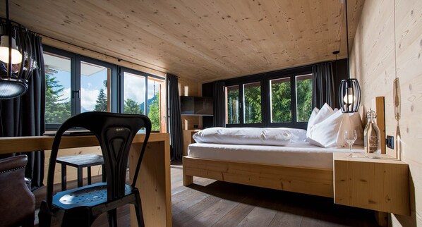 Una habitación con una cama de madera, un escritorio, una silla, ventanas amplias y vistas a árboles.
