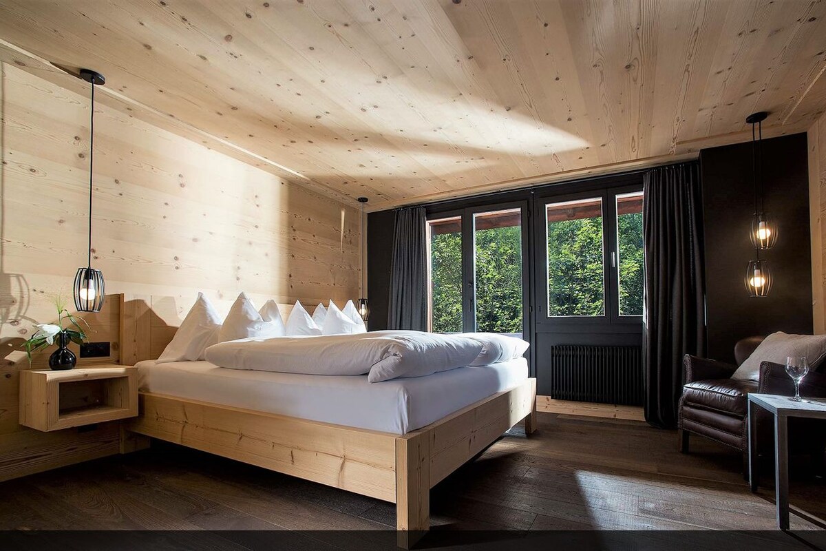 Un dormitorio con cama de madera, mesitas de noche, una mesita auxiliar, una ventana con cortinas, un radiador y una zona de estar con una silla y una mesa.