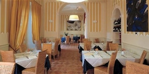 A restaurant with multiple tables and chairs, a chandelier, and a large painting on the wall.