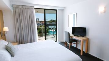 Habitación de hotel con una cama grande, un escritorio con televisor, una silla y vistas a una piscina y edificios.