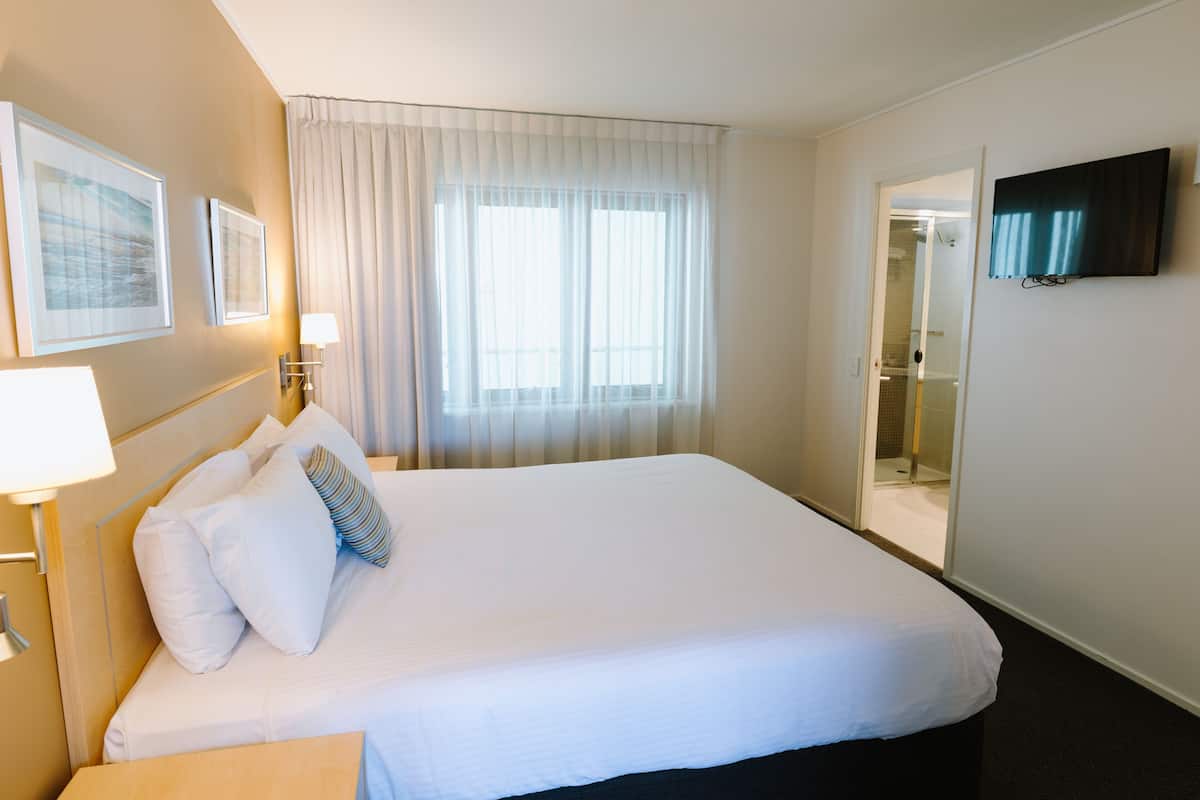 Ein Hotelzimmer mit einem großen Bett, einem Fernseher, einem Badezimmer mit Glastür und einem Fenster mit Vorhängen.
