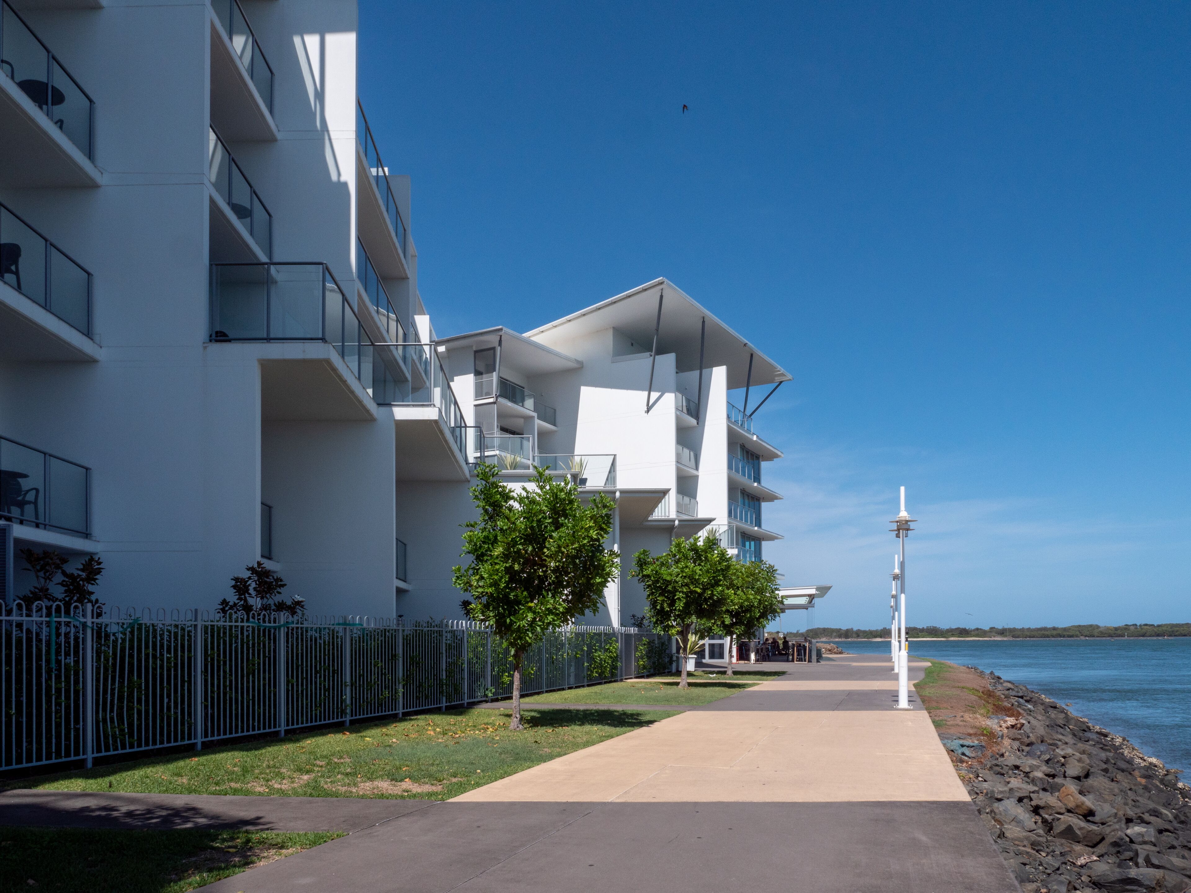 A modern multi-story building with balconies, a pathway leading to the water, and a clear view of the sea.