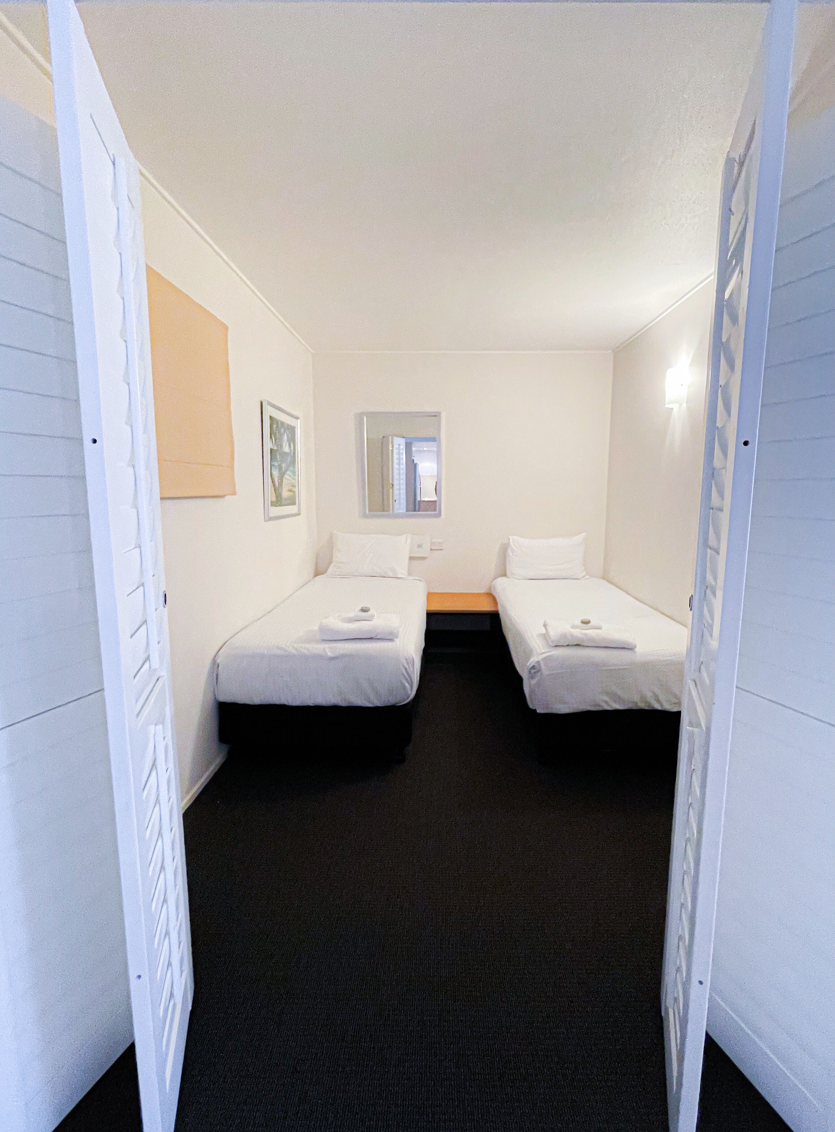 Une chambre d’hôtel avec deux lits simples, un petit miroir, un tableau encadré au mur et une moquette noire.