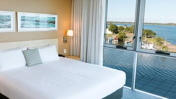 Habitación de hotel con una cama grande, una mesita de noche, una lámpara y vistas a un lago con barcos y muelles.