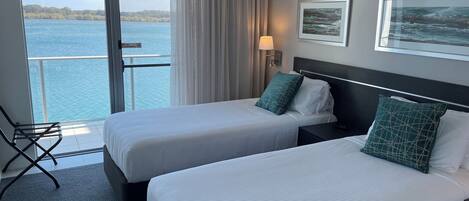 A hotel room with two beds, a view of the sea, a chair, and a lamp.