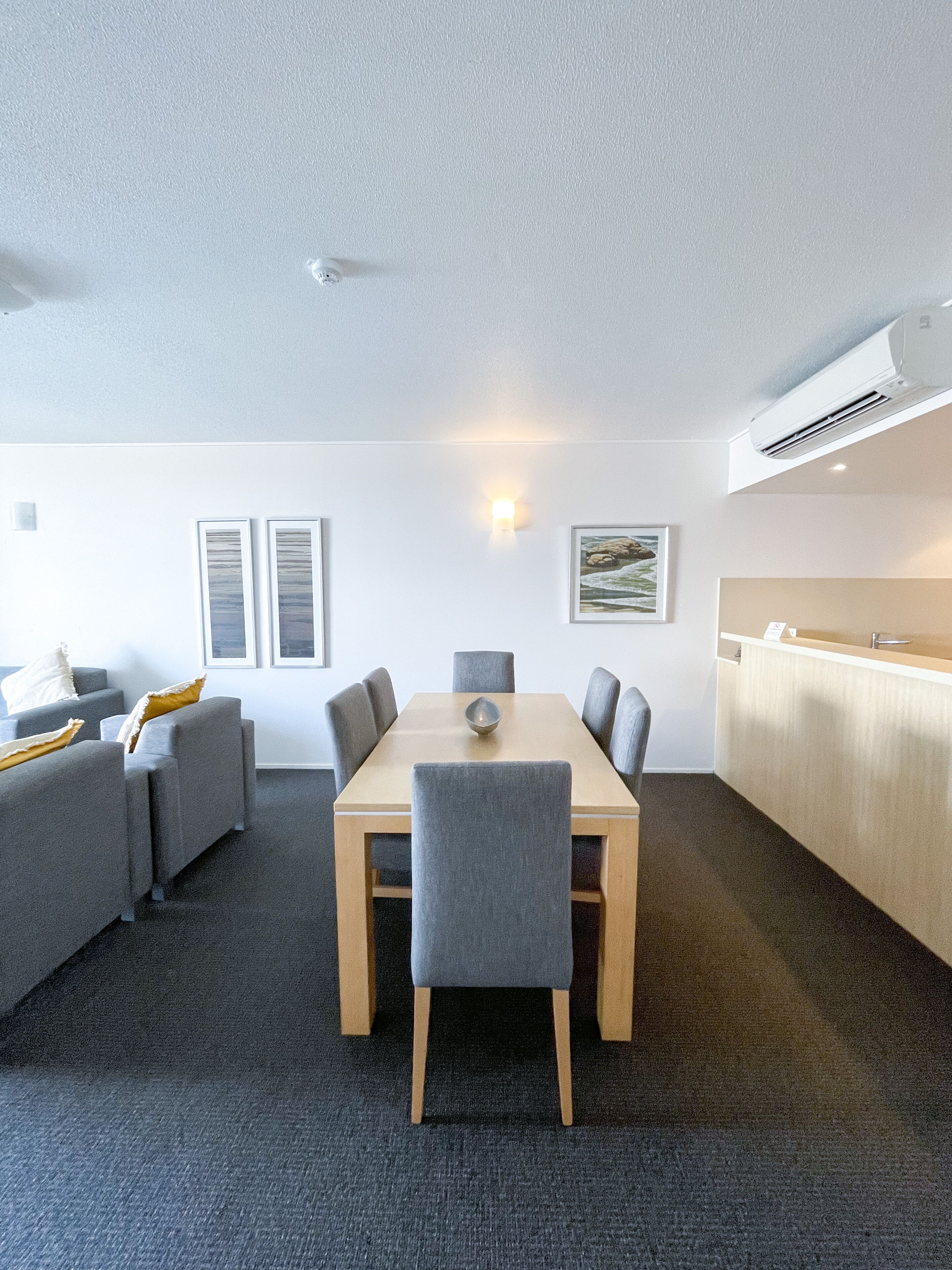 A modern room with a wooden dining table, grey upholstered chairs, a grey sofa, and a small kitchen area with a white countertop and an air conditioning unit.