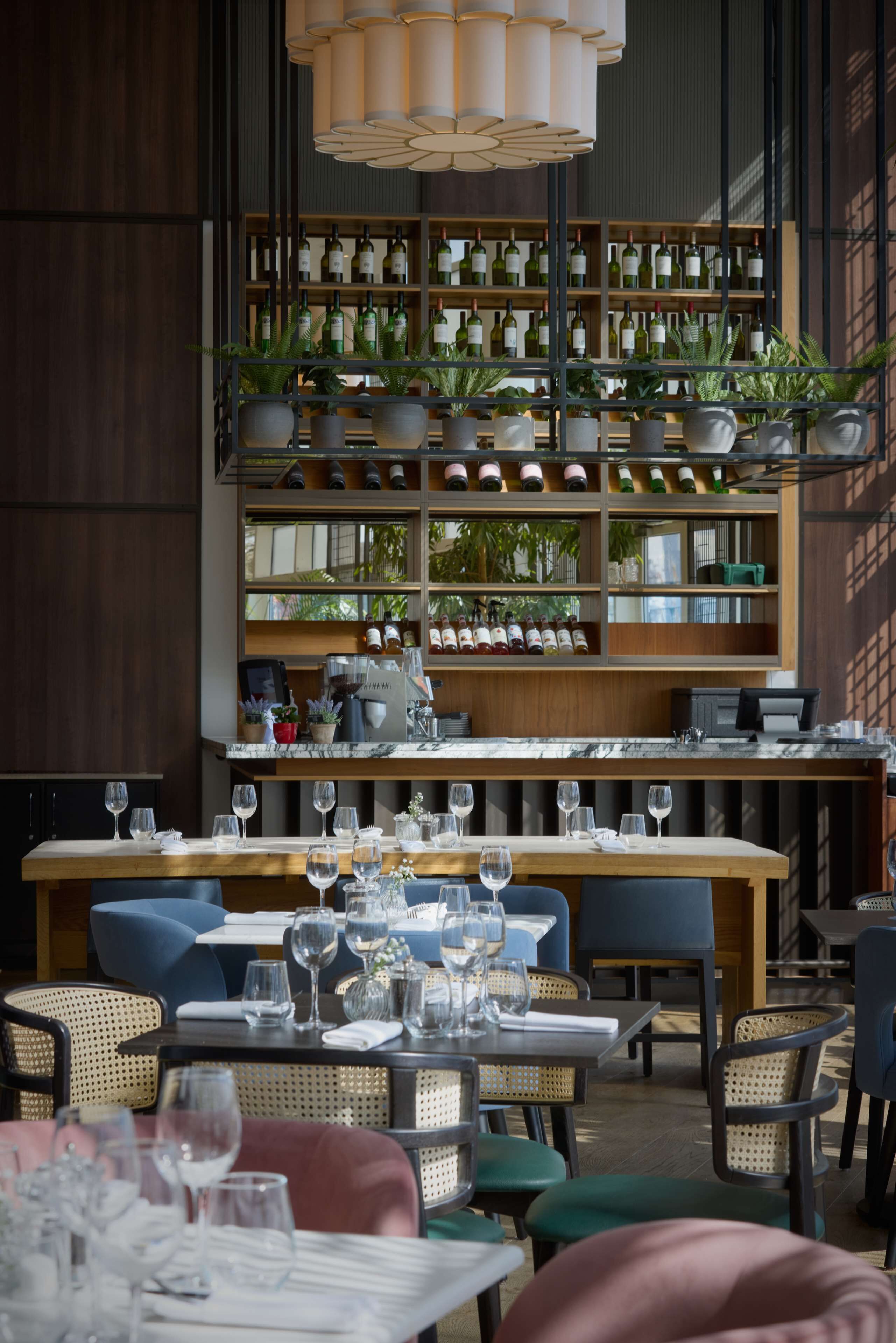 Ein Restaurantinneres mit einer langen Theke, mehreren Tischen und Stühlen. Auf Regalen stehen Weinflaschen und Blumentöpfe.
