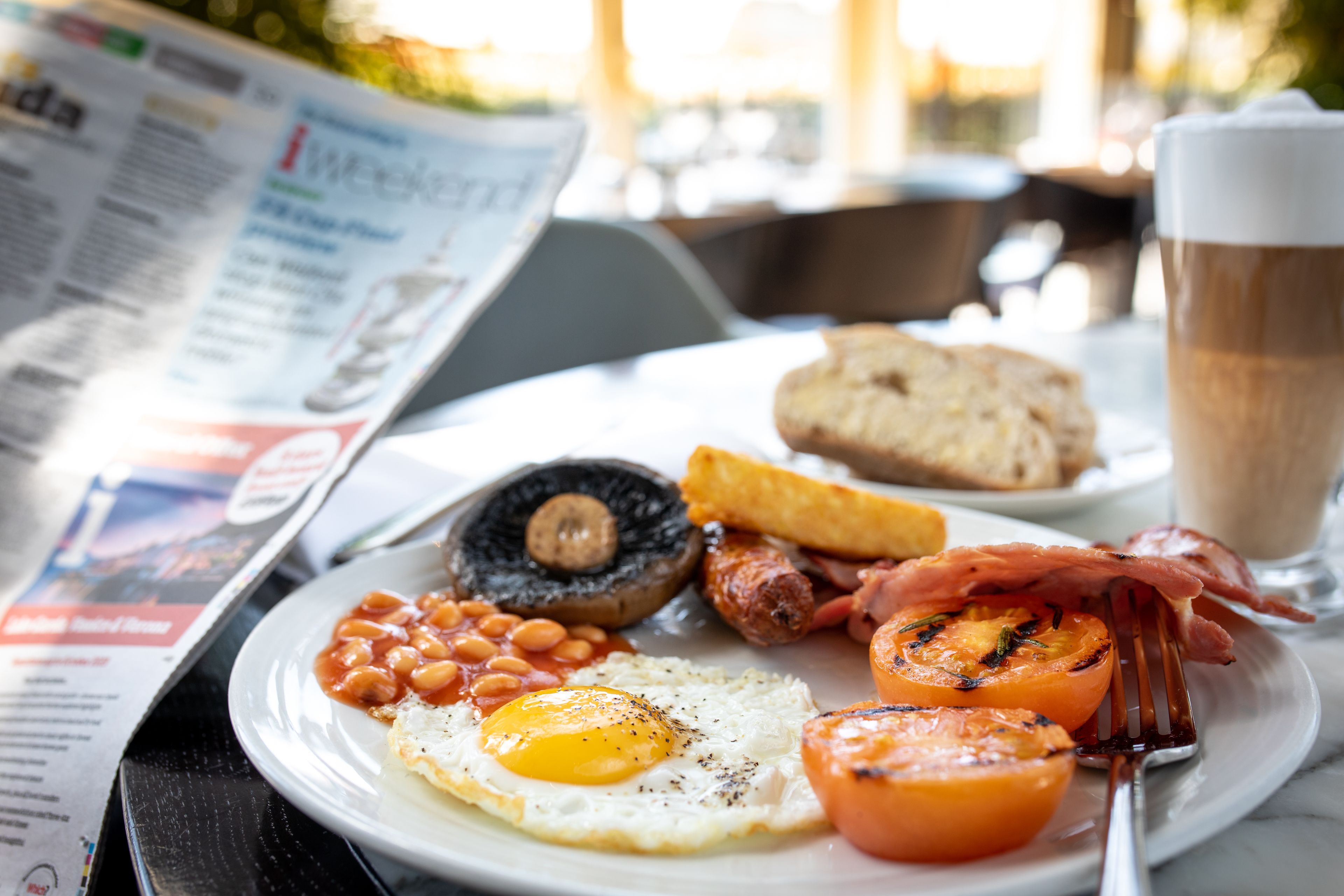 Un plato de desayuno con huevos, frijoles, champiñones, salchichas, tocino y tostadas, acompañado de una taza de café y un periódico.