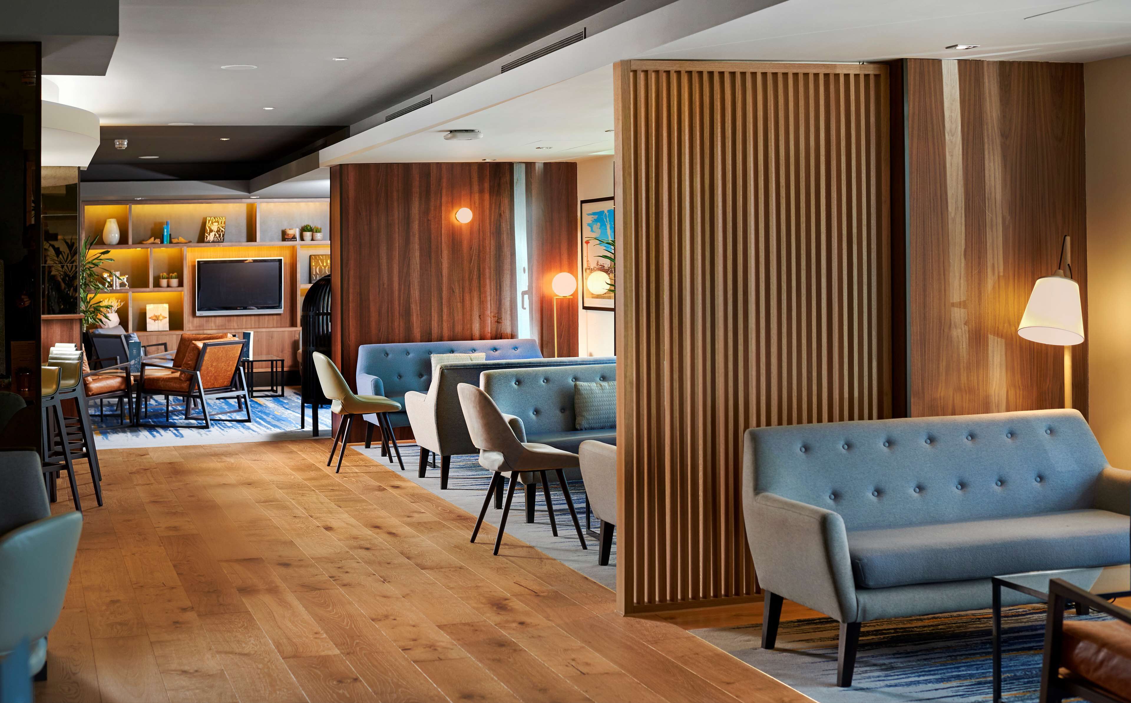 Un hall d’hôtel moderne doté d’un plancher en bois, d’espaces aménagés avec des sièges, d’un téléviseur à écran plat et d’un comptoir de réception.