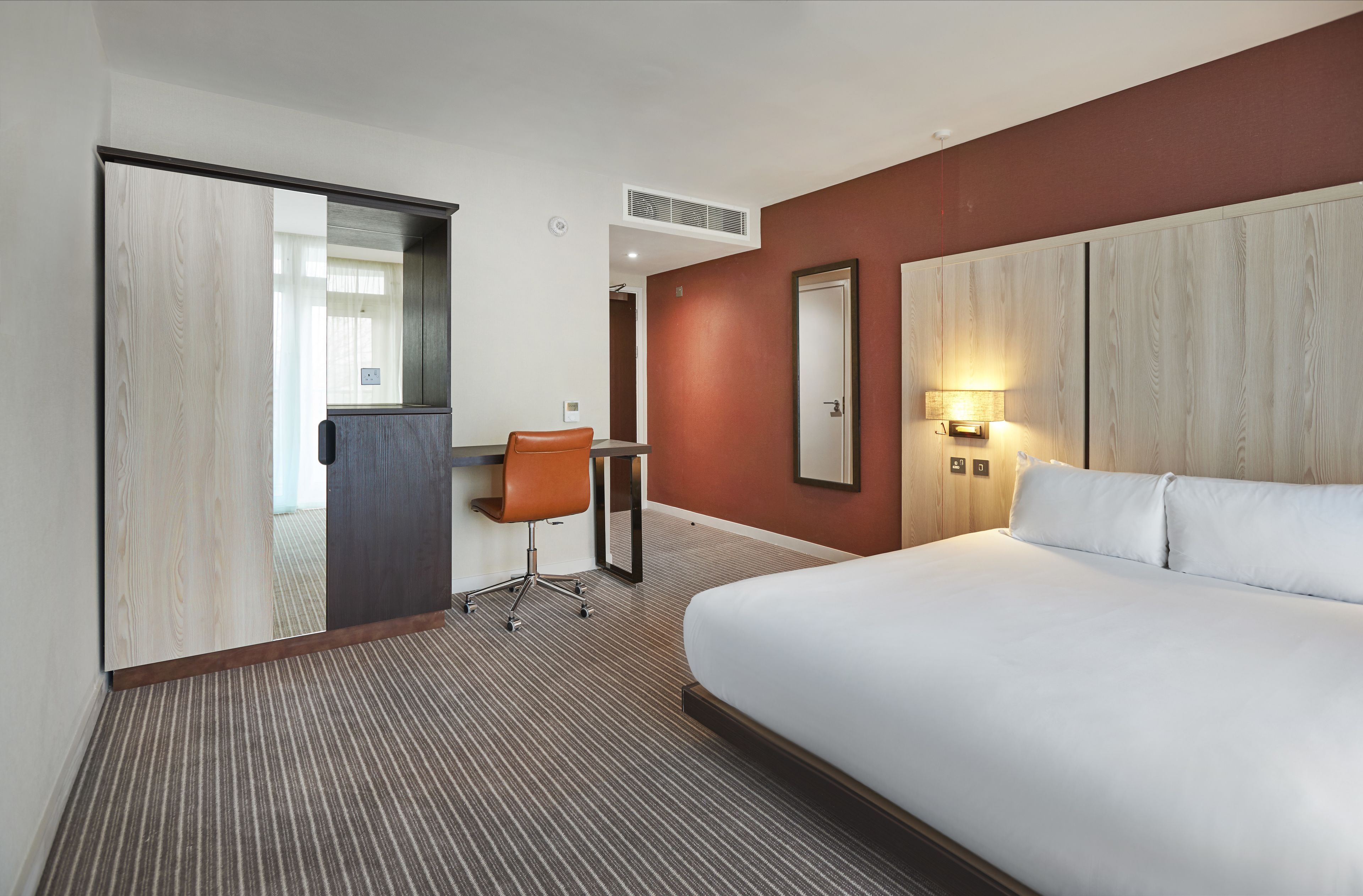 A hotel room with a large bed, a desk with a chair, a mirror, and a wall-mounted light fixture.