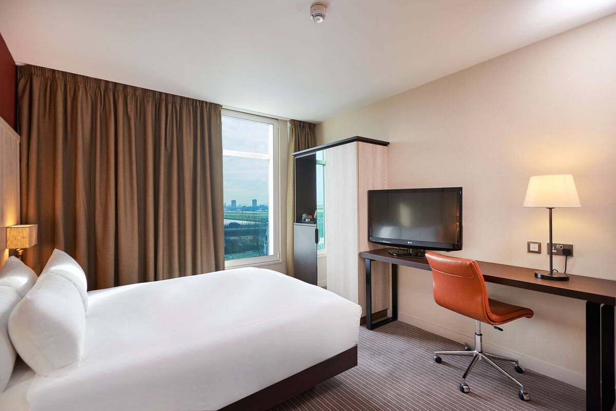 A hotel room with a bed, a desk with a chair, a television, and a view of the city through the window.