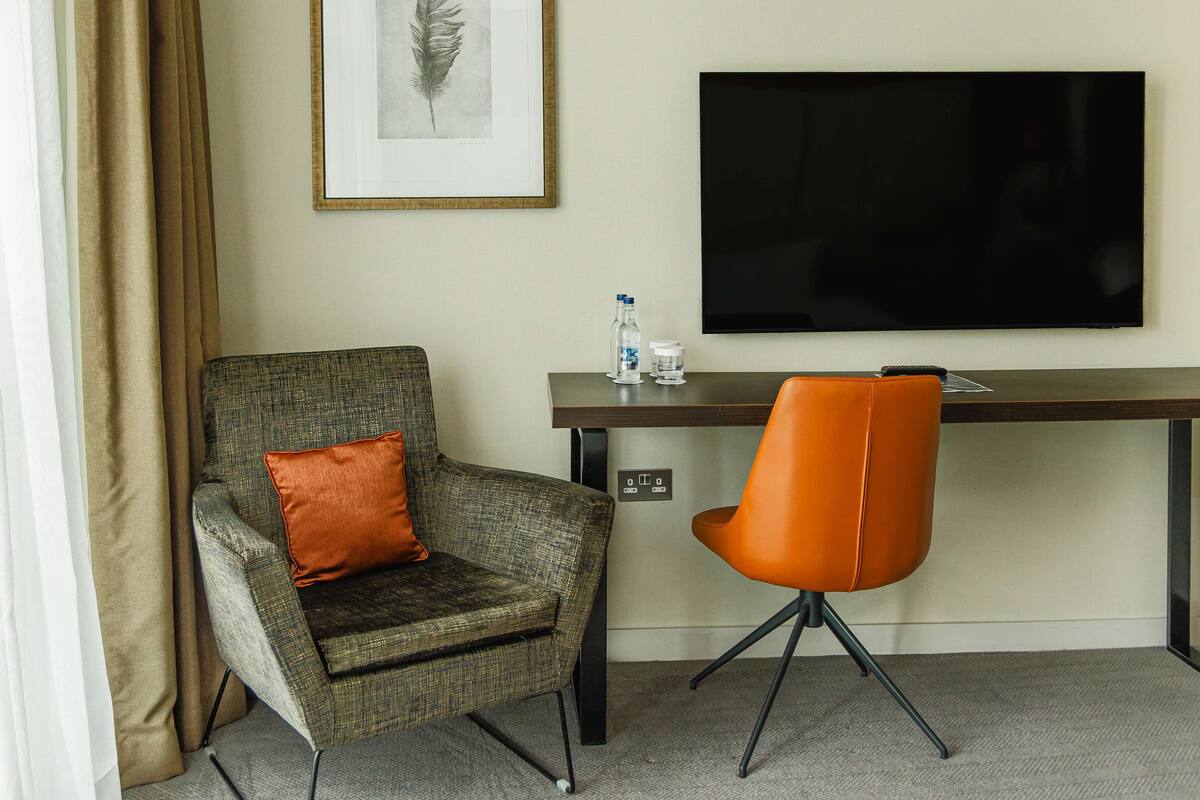A hotel room with a grey armchair, an orange chair, a flat-screen TV, a framed artwork, and a small table with a water bottle and a glass.