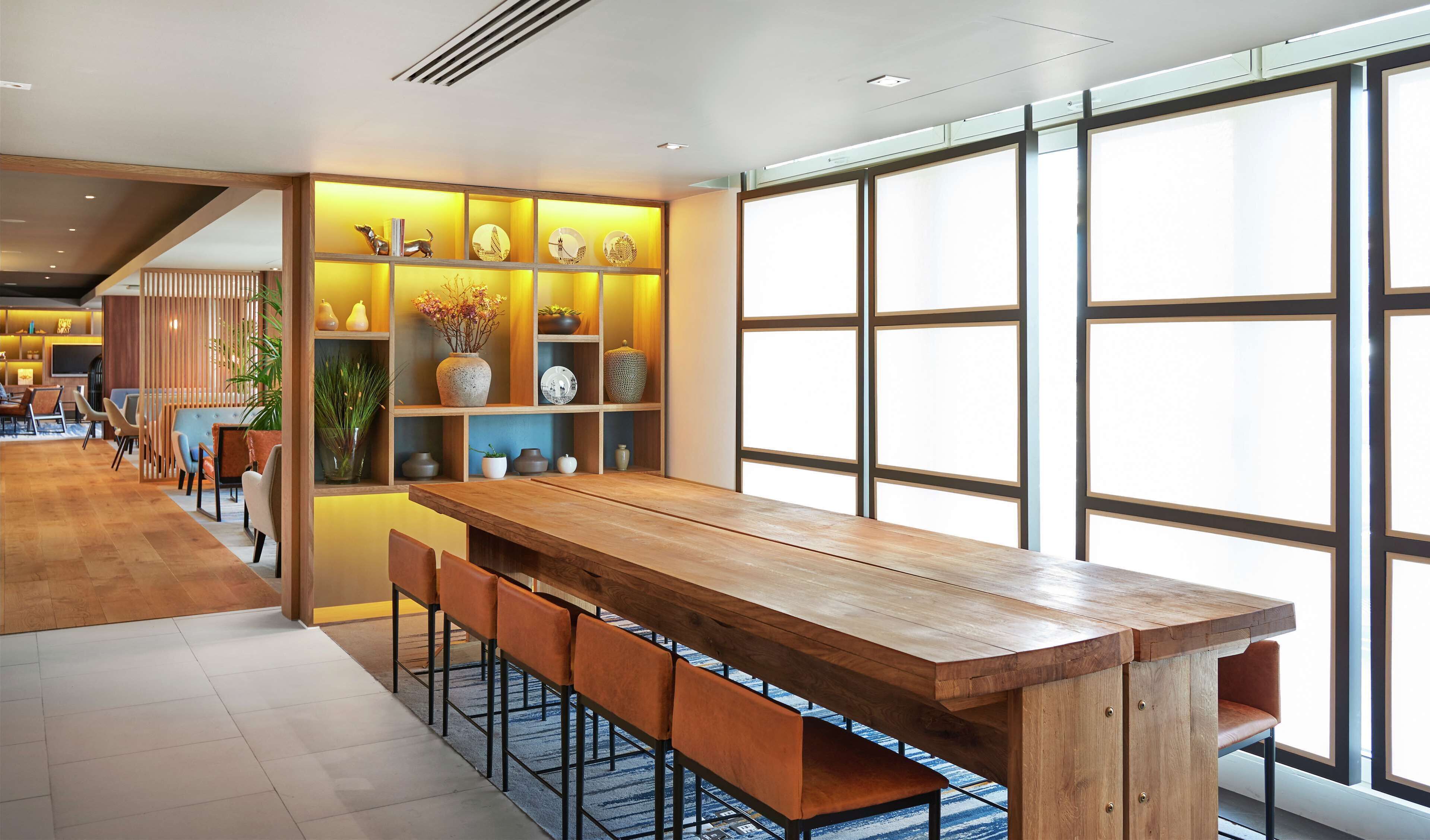 Un comedor moderno con una mesa de madera y taburetes de bar, una pared decorativa con estanterías y ventanas grandes que dejan entrar luz natural.