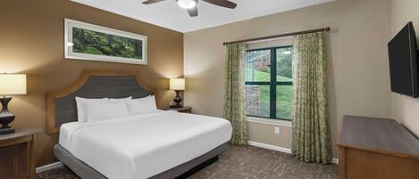 A hotel room with a bed, two bedside lamps, a ceiling fan, a window with curtains, and a mounted TV.