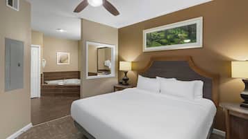 A hotel room with a large bed, a ceiling fan, a bathroom with a bathtub, and a framed picture of a forest on the wall.