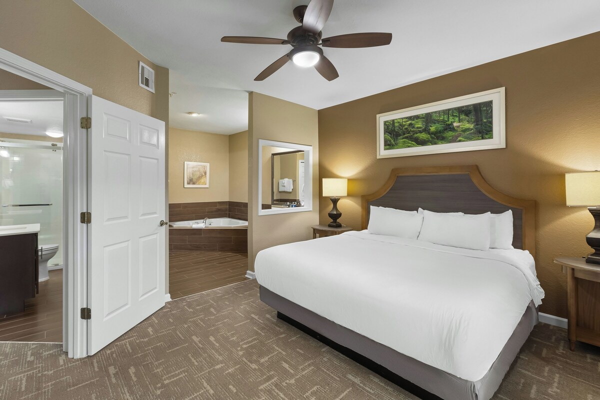 A hotel room with a bed, a bathroom, a ceiling fan, and a painting on the wall.