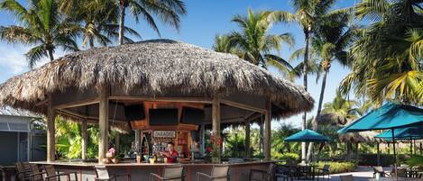 A thatched-roof bar area with outdoor seating and palm trees in the background.