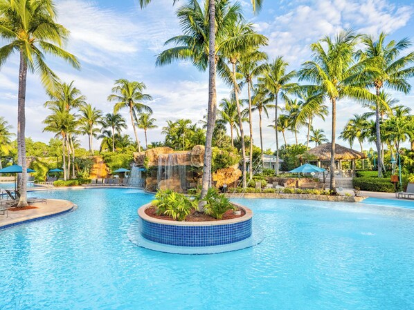 A tropical resort with a central pool, palm trees, a waterfall feature, and a thatched-roof structure.