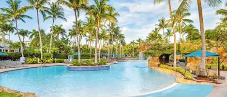 A large, clear swimming pool surrounded by palm trees and lounge chairs, with a rocky outcrop and a waterfall feature.