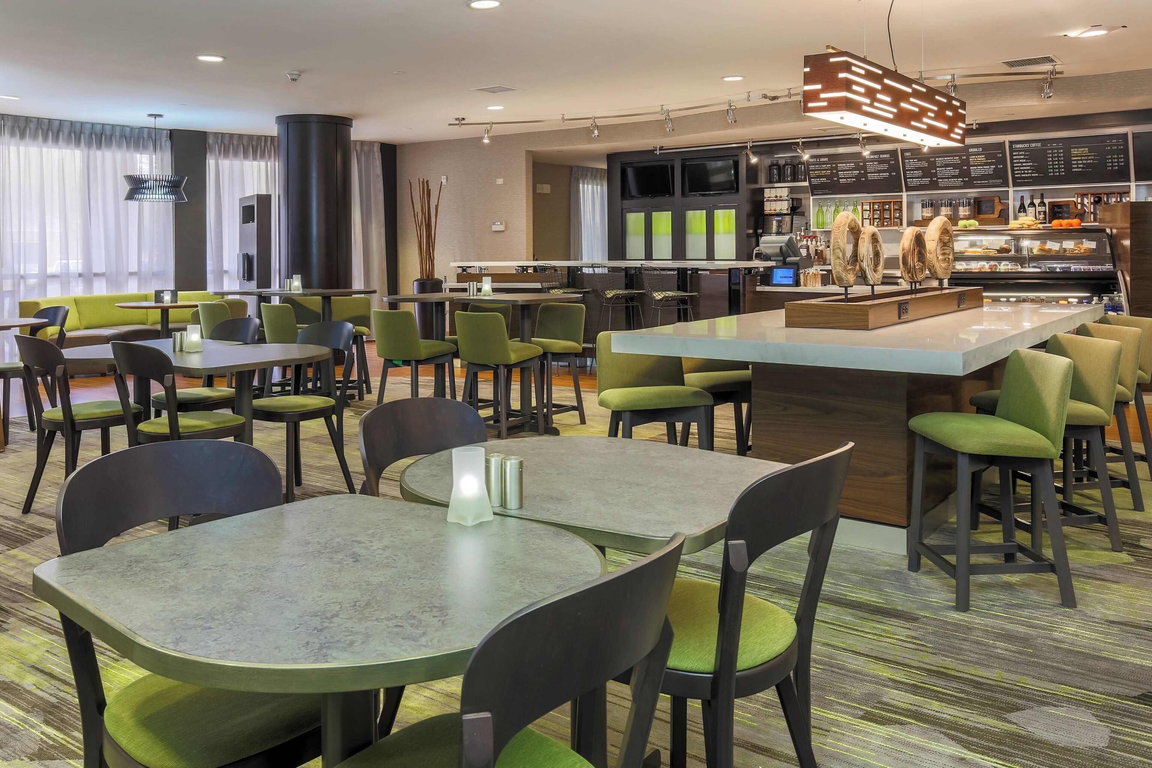 A modern restaurant with round tables and green chairs, a bar area with a counter, and a menu board in the background.