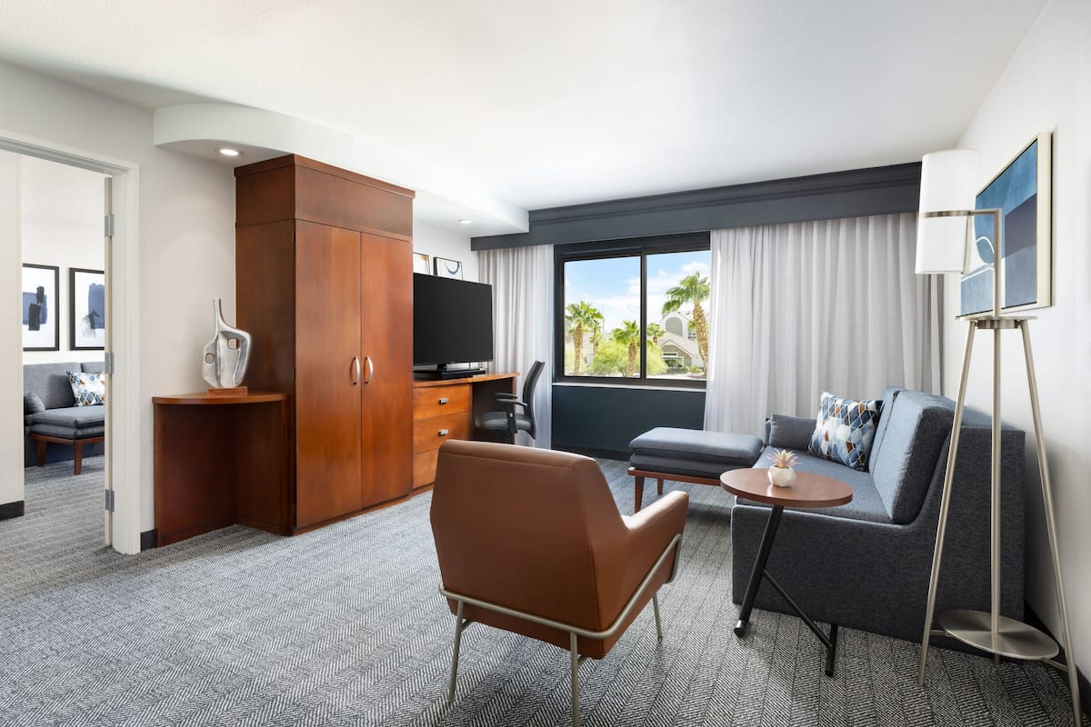 Ein modernes Hotelzimmer mit Sofa, Sessel, Fernseher und großem Fenster mit Blick auf Palmen.