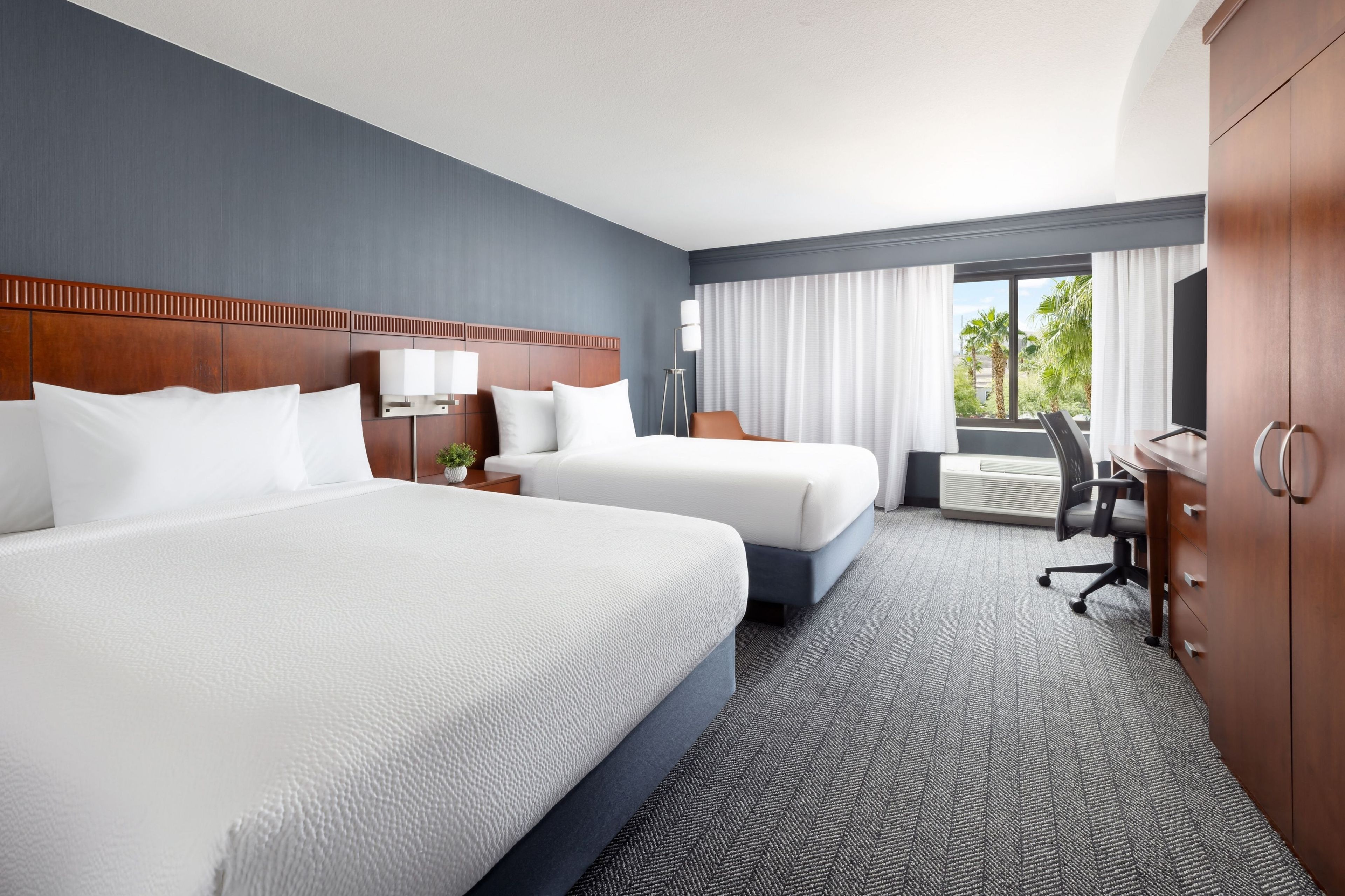 A hotel room with two beds, a desk, a chair, a wardrobe, and a large window with curtains.
