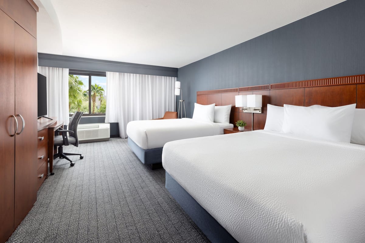 A hotel room with two beds, a desk, and a view of palm trees.