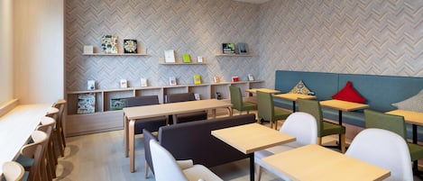 Un café moderno con mesas y sillas de madera, un revestimiento de pared con patrón de espiga y estantes con diversos objetos.