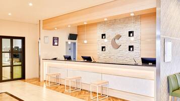 A reception area with a wooden reception desk, three bar stools, a stone wall with a large 'C' decoration, and a television mounted on the wall.
