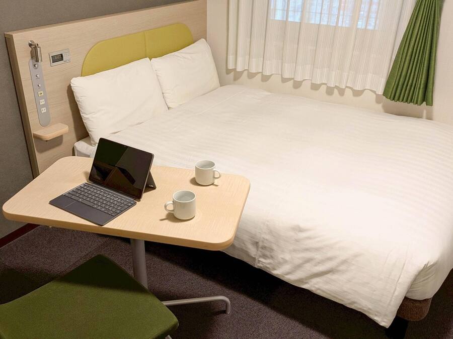 A hotel room with a bed, a desk with a laptop, two white mugs, and a green chair.