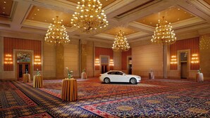 Un hall d’hôtel spacieux, agrémenté de lustres décoratifs, d’un tapis à motifs et d’une voiture blanche garée au centre.