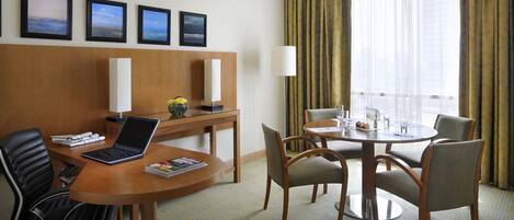 Una habitación de hotel con un escritorio de madera, una computadora portátil, una silla, una mesa de comedor con sillas, una lámpara y cuadros en la pared.
