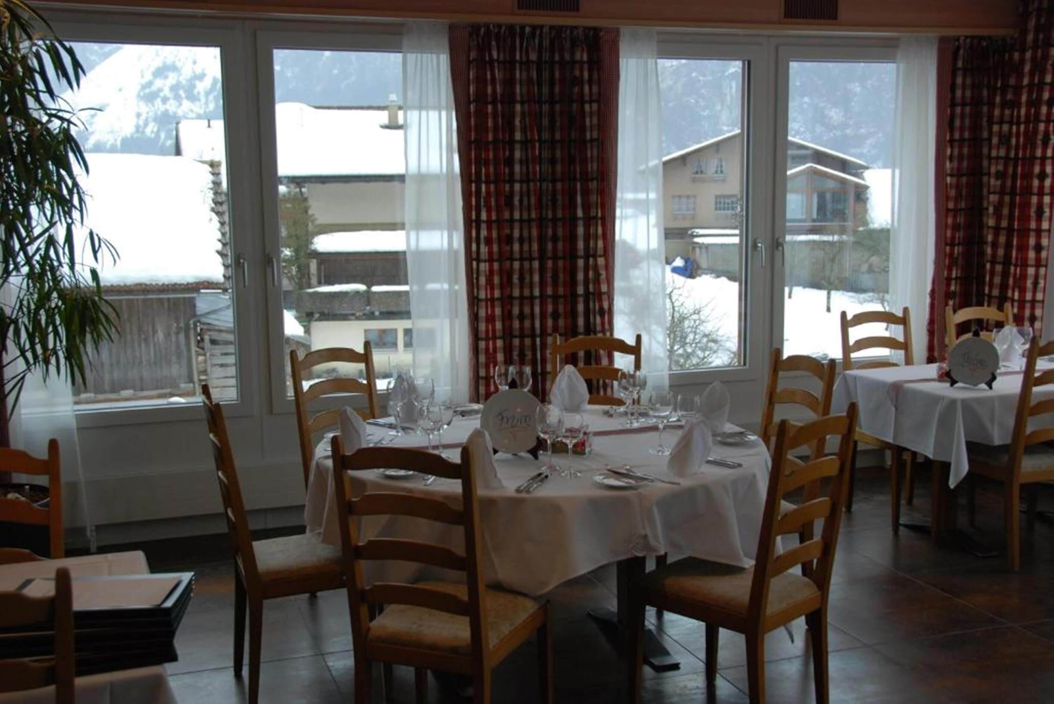 A dining area with tables set for four, wooden chairs, and a view of snow-covered buildings through large windows.