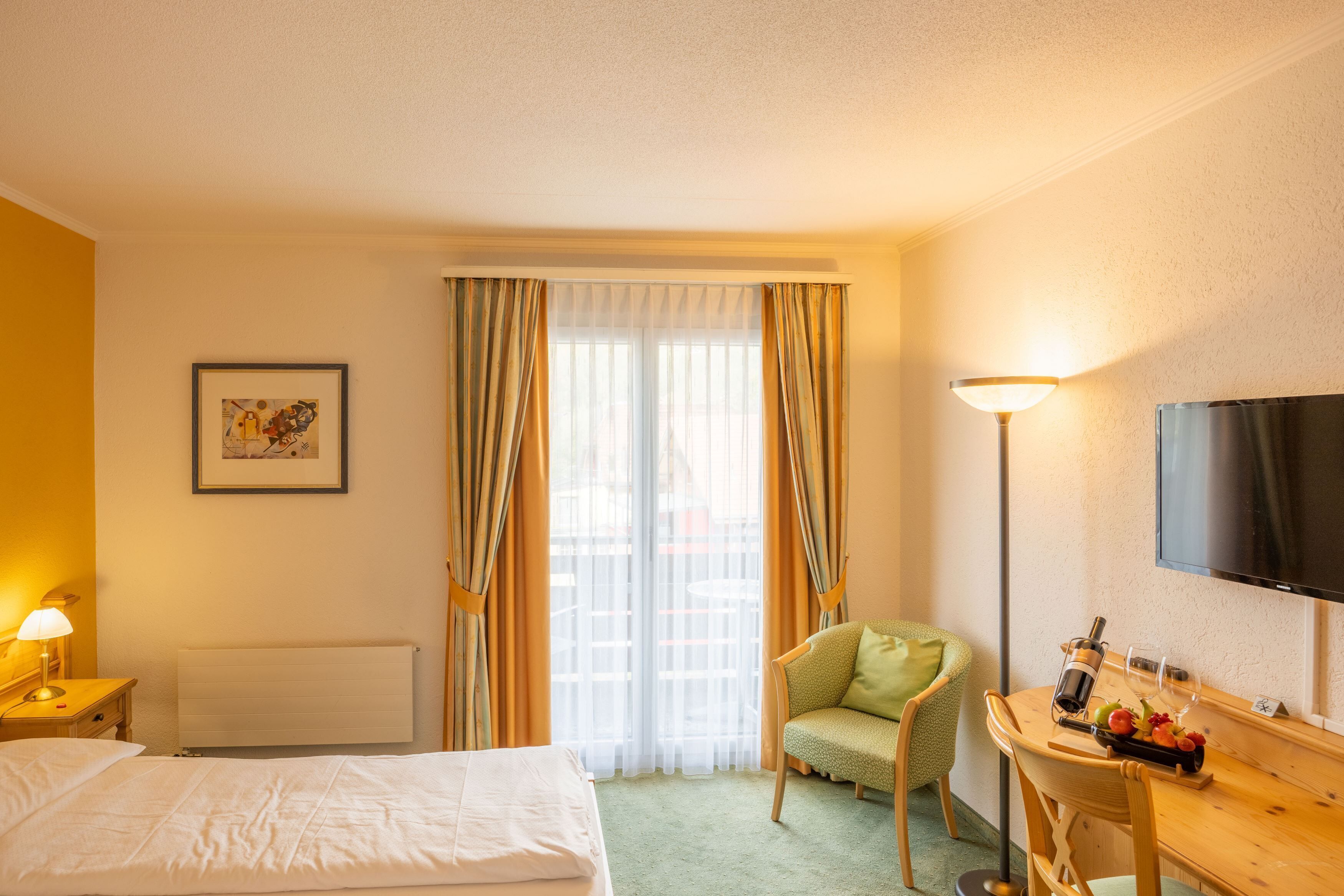 Habitación de hotel con cama, una silla, escritorio con una botella y vasos, televisión y ventana con cortinas.
