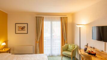 Habitación de hotel con cama, una silla, escritorio con una botella y vasos, televisión y ventana con cortinas.