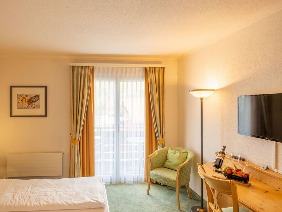 Habitación de hotel con cama, una silla, escritorio con una botella y vasos, televisión y ventana con cortinas.